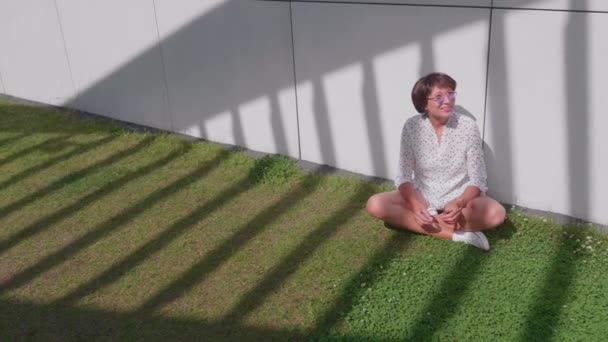 Frau mit bunter Sonnenbrille sitzt auf Rasen im Stadtpark und genießt das helle Sonnenlicht. Sommerhitze in der Stadt. — Stockvideo