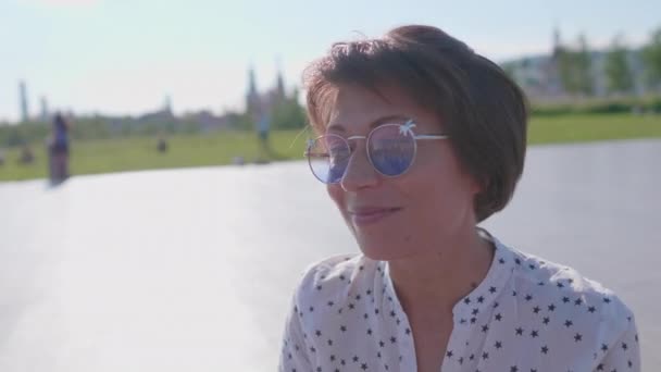 Woman with colorful sunglasses sits on scene in urban park and enjoys of bright sunlight. Summer heat in town. — Stock Video