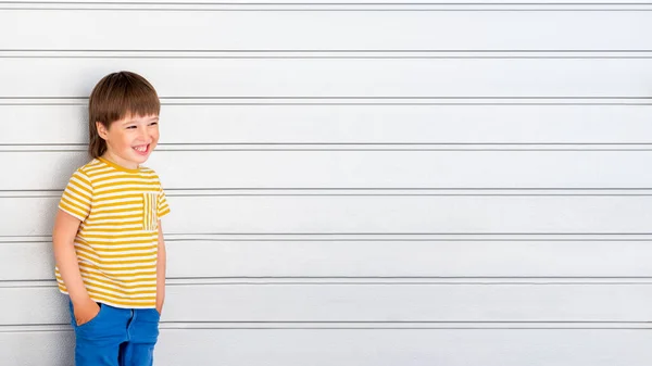 一个有着条纹的白色背景的笑男孩的画像 可爱的孩子手插在浅灰色的墙边的口袋里 穿着黄色T恤和蓝色牛仔裤的孩子笑得很开心 — 图库照片