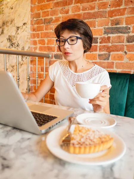 Woman works remotely in cafe with red brick walls. Female in eyeglasses types on laptop keyboard, drinks coffee and eats a tart. Co-working center with loft interior style. Freelance workplace.