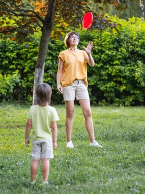 Anne ve oğlu çimlerin üzerinde frizbi oynuyorlar. Yaz titreşimleri. Açık hava eğlence aktivitesi. Aile hayatı. Arka bahçede spor maçı.