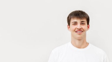 Kısa saçlı ve metal telli yakışıklı bir genç. Açık gri arka planda beyaz tişörtlü adamın portresi. 