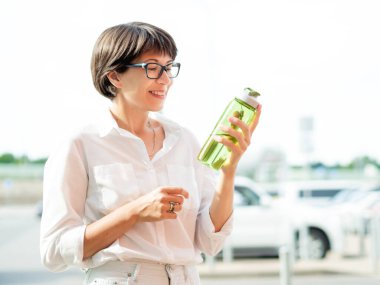 Susamış kadın doldurulmuş yeşil şişeden su içiyor. Sıvılar için kullanışlı şeffaf bir şişe. Sıcak yaz havası.