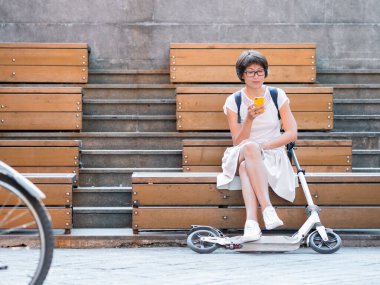 Kadın tahta bankta oturuyor ve scooter 'a bindikten sonra akıllı telefonuna mesaj atıyor. Sağlıklı bir yaşam tarzı. Çevre dostu şehir ulaşımı. 