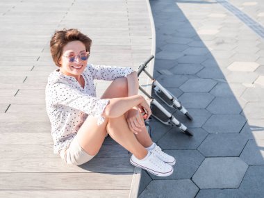 Rüzgar, renkli güneş gözlüklü çilli bir kadının kısa saçlarını dalgalandırıyor. Açık ahşap sahnede gülümseyen kadın. Yaz titreşimleri. Samimi duygular. Kick scooter - Çevre dostu şehir taşımacılığı.