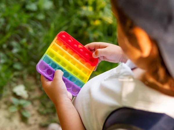 Little boy plays square rubber fidget toy Pop It. Rainbow-colored anti stress and tactile toy for all ages. Outdoor recreation with toy for fine motor skills improvement.