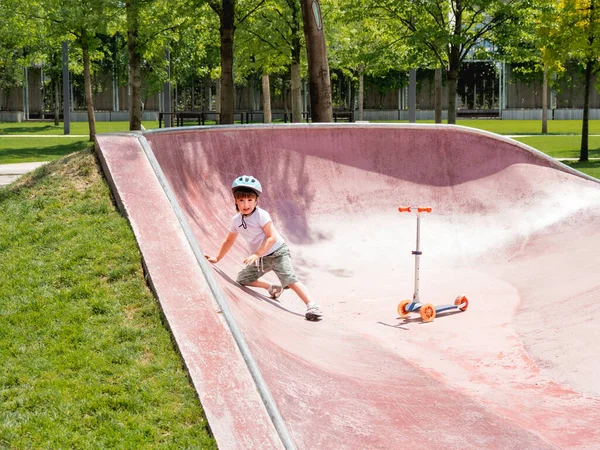 Little boy fell off kick scooter while riding in skate park. Special concrete bowl structures in urban park. Training to skate at summer.