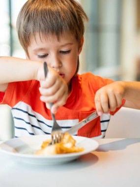 Kırmızı tişörtlü şirin çocuk çatal ve bıçakla krep yiyor. Yüzünde komik bir ifade olan meraklı bir çocuk. Kahvaltı için lezzetli hamur işi..