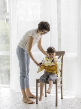Anne oğlunun saçını tek başına kesti. Küçük çocuk kumaşla kaplı bir şekilde oturur ve elinde bir makas tutar. Koronavirüs COVID-19 karantina ve tecrit durumunda yeni normaller. Ev yaşamında güzellik.