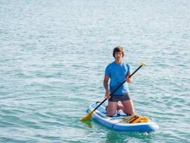 Kürekçi. Sporcu dizlerinin üzerinde ayakta kürek çekiyor. Sup sörfü. Aktif yaşam tarzı. Açık hava eğlencesi. Deniz kenarında tatil..