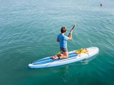 Kürekçi. Sporcu dizlerinin üzerinde ayakta kürek çekiyor. Sup sörfü. Aktif yaşam tarzı. Açık hava eğlencesi. Deniz kenarında tatil..
