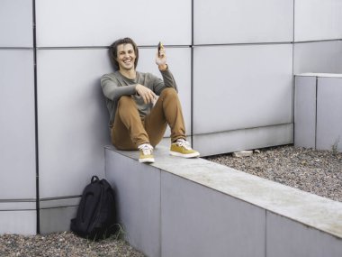 Gülen adam akıllı telefonuyla duvarın dışında oturuyor. Sırt çantalı, sıradan giyinmiş bir öğrenci. Uzun saçlı, yakışıklı bir adam. Modern şehir yaşam tarzı.