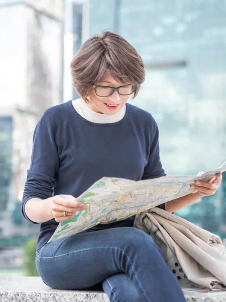 Smiling woman in eyeglasses is reading paper map sitting on background of buildings with glass walls. Travel around city. Urban tourism. Modern architectural landmarks.