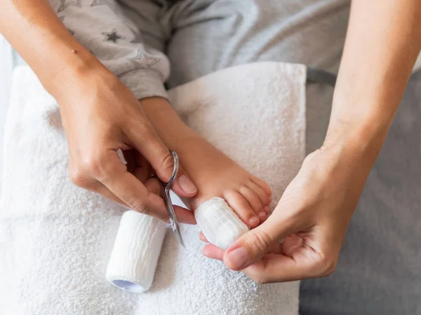 Mother bandages her child\'s big toe. Close-up photo of kid\'s foot with bandaged finger. First aid in case of small domestic injury.