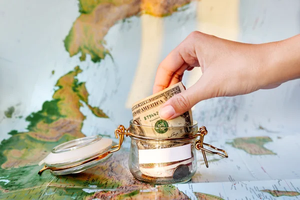 Collecting money for travel. Glass tin as moneybox with cash savings (banknotes and coins) on map as background. White empty space for your text. Moneybox in sunlight. Woman puts one dollar banknote in jar. — Stock Photo, Image