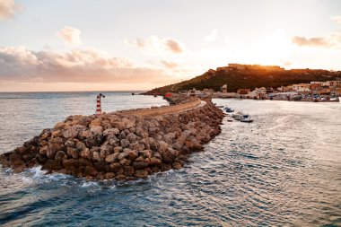 Entrance in bay, seafront with lighthouse on seafront at sunset. Gozo, Malta.