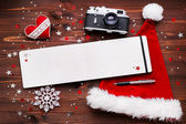 Christmas and New Year background with old fashioned camera, red Santas hat, notepad with pen and christmas decorations - stars, silver sparkling snowflakes, confetti on wooden table.