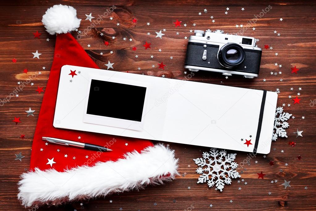 Christmas and New Year background with old fashioned camera, red Santa's hat, notepad with pen, empty photo frame and christmas decorations - stars, silver sparkling snowflakes, confetti on wooden table. Place for your text.