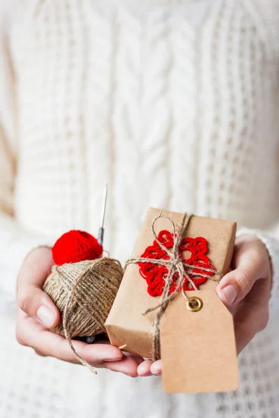 Woman in white knitted sweater and mitts holding a present and coils of red thread and linen rope. Gift is packed in craft paper with hand made crocheted red snowflake. Present has an empty tag for your text. Mock up. Example of DIY ways to pack Cris — Stock Photo, Image