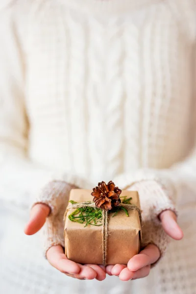 Woman in knitted sweater holding a present. Gift is packed in craft paper with pine cones and tied with rough rope. Example of DIY way to wrap a present. Place for your text.