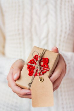 Örme kazak bir hediye tutan kadın. Hediye Kraft kağıt ile el yapımı tığ işi kırmızı kar tanesi doludur. Hediye metniniz için boş bir etiketi var. Kadar alay et. Noel hediye paketi için DIY yolu