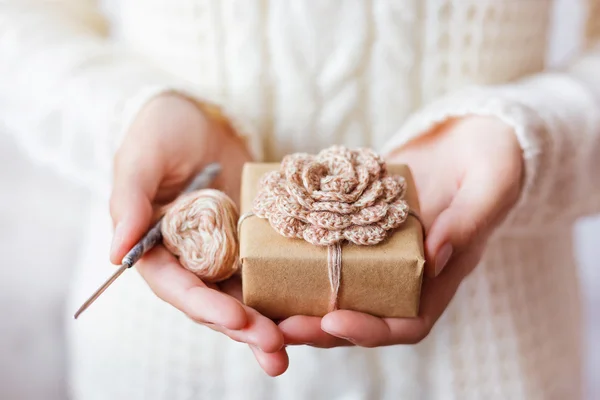 Woman in white knitted sweater holding a present and coil of thread with crochet hook. Gift is packed in craft paper with hand made crocheted flower. — Stock fotografie