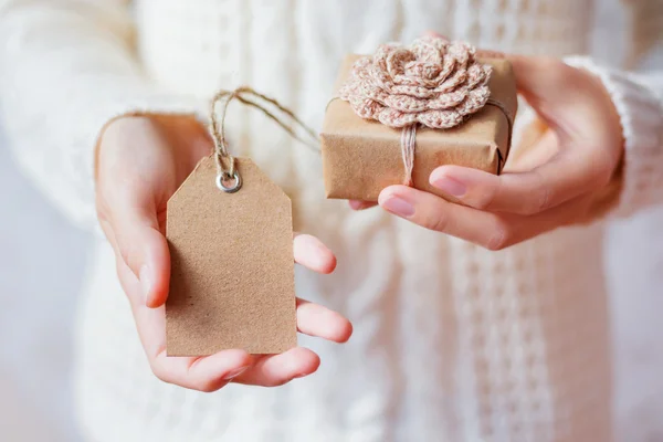 Woman in white knitted sweater holding a present. Gift is packed in craft paper with hand made crocheted flower.Empty tag for your text.