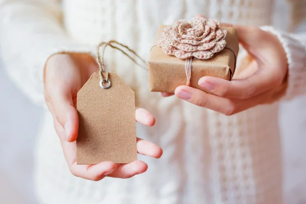 Mujer en jersey de punto blanco sosteniendo un regalo. El regalo se embala en papel artesanal con ganchillo hecho a mano flower.Empty etiqueta para su texto . —  Fotos de Stock