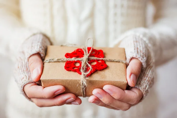Donna in maglia bianca maglione e guanti con in mano un regalo. Il regalo è confezionato in carta artigianale con fiocco di neve rosso all'uncinetto. Esempio di modo fai da te per avvolgere un regalo . — Foto Stock