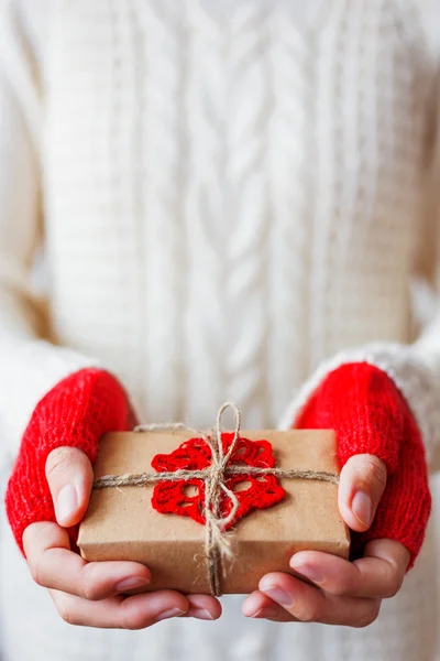 Mulher de camisola de malha branca e luvas segurando um presente. Presente é embalado em papel artesanal com floco de neve vermelho de malha. Exemplo de forma DIY para embrulhar um presente . — Fotografia de Stock