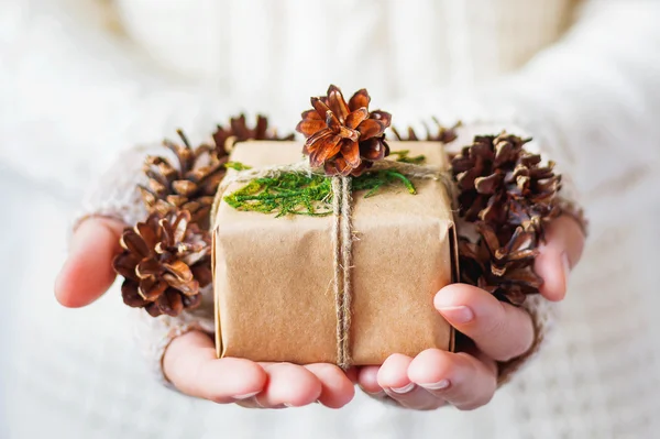 Woman in knitted sweater holding a present. Gift is packed in craft paper with pine cones and tied with rough rope. Hands full of pine cones.