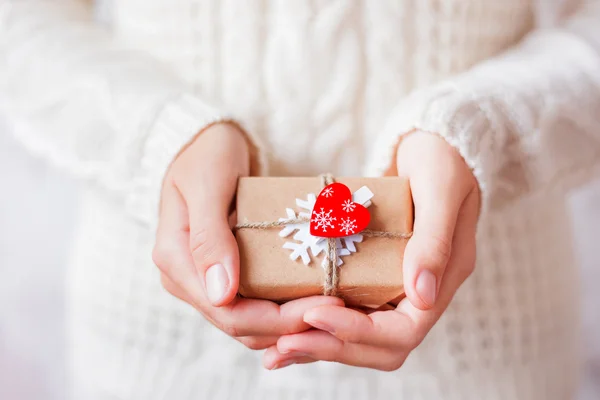 Donna in maglia con un regalo in mano. Il regalo è confezionato in carta artigianale con fiocco di neve bianco e cuore rosso . — Foto Stock