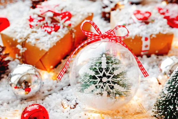 Weihnachten und Neujahr Hintergrund mit Geschenken, Bändern, Kugeln und verschiedenen Dekorationen auf Schnee. transparente Kugel mit Schneeflocke und Tanne im Inneren. — Stockfoto