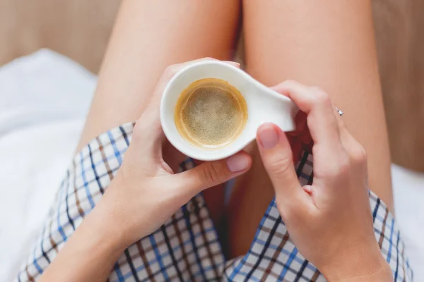 Vrouw in shirt zit op bed met een kop met warme koffie. — Stockfoto