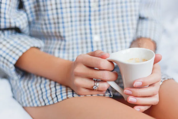 Vrouw in shirt zit op bed met een kop met warme koffie. — Stockfoto