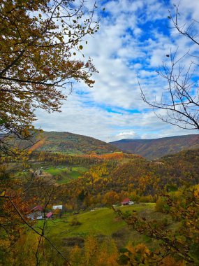 Sonbahar mevsiminde dağlardaki küçük köyün güzel manzarası. Etrafı renkli bir orman ve bulutlu mavi bir gökyüzü. Muntele Rece, Cluj, Romanya