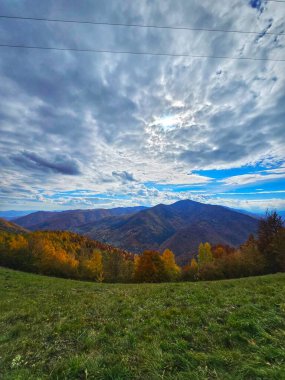 Dağın tepesinden yeşil otlaklı güzel manzara, sarı, turuncu ve kırmızı yapraklı ormanlar sonbahar gününde mavi gökyüzünde beyaz bulutlar. Muntele Rece, Cluj, Romanya