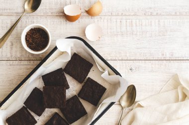 Fırın kağıdının içinde browniler, yumurta kabukları, kasede çikolata, iki kaşık ve beyaz peçete, beyaz ahşap bir fon üzerinde.. 