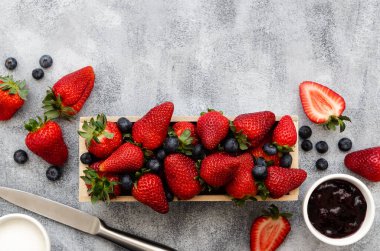 Çilek ve yaban mersini ahşap bir dikdörtgen kutuda, çilek ve yaban mersini reçeli ve şeker beyaz kaselerde bıçakla, gri arka planda. 