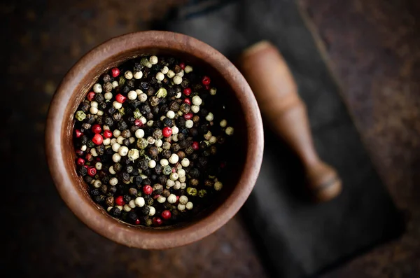 Black, white, green, and pink peppercorns in a wooden mortar with pestle on a brown napkin and dark backdrop.