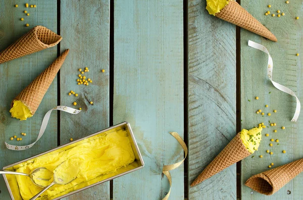 Peach ice cream in a silver container and in cones, with yellow candies and golden and white ribbons on light blue wooden backdrop.