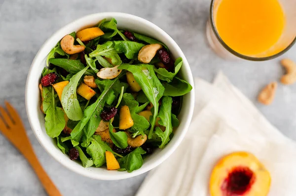 Arugula, sarı şeftali, kaju fıstığı, ve beyaz kasede kurutulmuş kızılcık salatası, ahşap çatal, bir bardak portakal suyu ve yarım şeftali beyaz peçete ve gri fon.. 