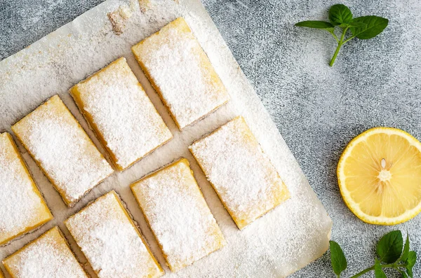 Lemon squares on baking paper, a slice of lemon and mint leaves, on a grey backdrop.