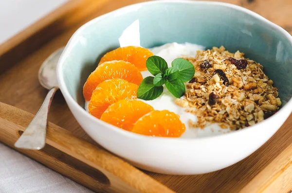 Sade yoğurt, mandalina segmentleri, granola, ve nane yaprakları beyaz ve açık mavi bir kasede, bir kaşıkla, ahşap tepside. 