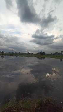 Dramatik bulutlu gökyüzünün altında, doğa tarafından çevrelenmiş, su yüzeyindeki yansımalarla sakin bir kırsal alan yaratan, su kültürünün huzurlu bir görüntüsü..