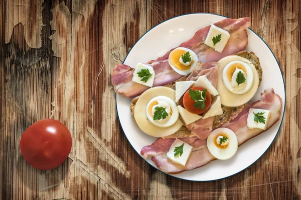 Teller mit Speck-Käse-Eierschinken und Tomaten-Sandwich auf altem Holzgrund — Stockfoto