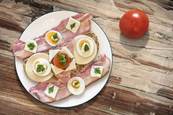 Teller mit Speck-Käse-Eierschinken und Tomaten-Sandwich auf altem Holzgrund — Stockfoto