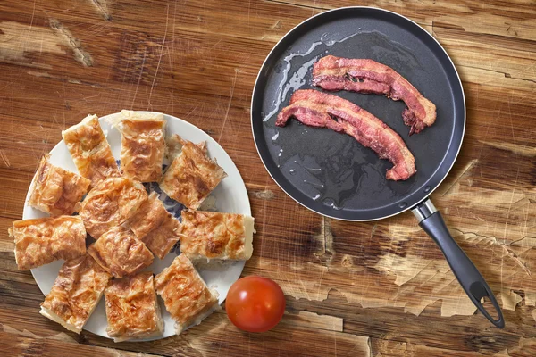 Arandelas de tocino frito en sartén con plato de rodajas de pastel de queso Gibanica y tomate en mesa de madera vieja — Foto de Stock