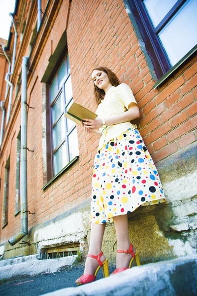 美丽的女孩在黄色的浪漫礼服与书附近的老式红砖墙 — 图库照片