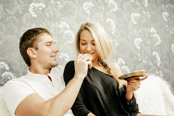 年轻的白种白人夫妇坐在白色的羊毛沙发上一起吃饼干的肖像 — 图库照片